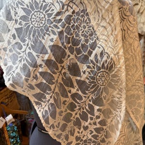 May include: A sheer, beige scarf with a large floral pattern in a darker gray. The design features stylized flowers and leaves. The scarf is draped over a mannequin, showcasing its flowing fabric and intricate print.