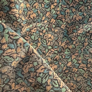 May include: Close-up of a fabric with a repeating leaf pattern. The leaves are in shades of green, brown, and gray against a dark background. The fabric is folded, revealing the texture and pattern.