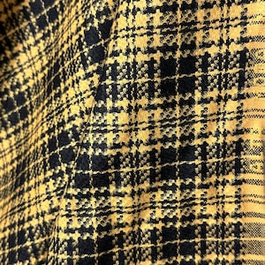 May include: Close-up of a fabric with a houndstooth pattern. The fabric features a repeating design of black and yellow squares and rectangles. The fabric is folded, creating shadows and texture.