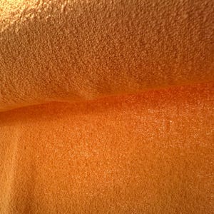 May include: Close-up of a rolled-up, textured orange fabric. The fabric has a soft, plush appearance with a slightly uneven surface. The color is a vibrant, warm orange, with subtle variations in tone across the surface.