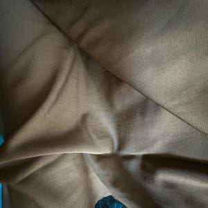 May include: A solid brown fabric with a fine, woven texture and visible diagonal folds. The material appears opaque and smooth, with subtle variations in shading across the surface due to the draping, creating a soft, matte appearance throughout.