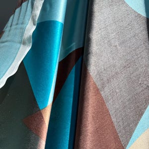 May include: Close-up of a fabric with a geometric pattern. The design features large shapes in shades of teal, brown, gray, and light blue. The fabric appears to be silky and has a slight sheen, with folds and shadows adding depth.