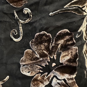 May include: A black fabric featuring a raised, textured floral pattern in a dark brown velvet finish. The design includes large, detailed flower blossoms and delicate, swirling vine-like stems that create a tactile, dimensional surface.