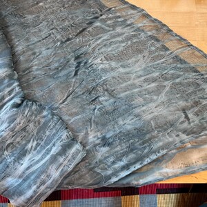 May include: A draped, sheer scarf in shades of gray and blue with a textured, marbled appearance. The fabric is folded and spread out on a wooden surface, showcasing its flowing design. The scarf's edges are pointed.