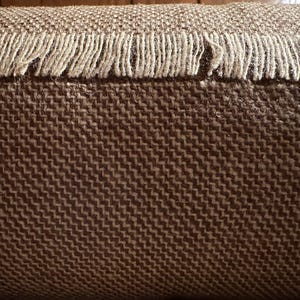 May include: Close-up of a brown woven fabric with a fringe border. The fabric has a subtle herringbone pattern.