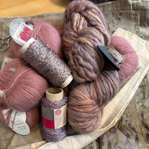 May include: A collection of yarn supplies including several skeins of dusty rose wool yarn, two spools of metallic thread in shades of bronze and purple, and a pair of silver scissors, all arranged together on a textured beige fabric surface.