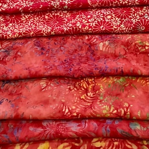 May include: A stack of red fabric with various floral patterns. The top layers feature a repeating white floral design on a red background. The lower layers display a batik-style pattern with red, purple, and yellow floral motifs.