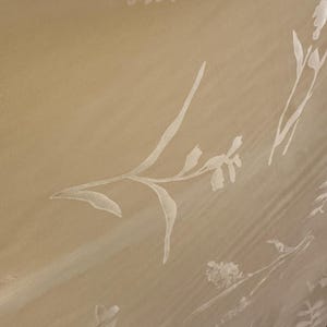 May include: A sheer, cream-colored fabric with a delicate floral pattern. The design features stylized white flowers and leaves, creating a subtle, elegant texture. The fabric appears to be draped, with soft folds and shadows.