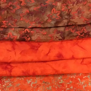 May include: Stack of four fabric squares in various shades of orange and brown with different patterns. The top square is a brown and orange floral pattern, the second square is a solid orange, the third square is a light orange with a white floral pattern, and the bottom square is a darker orange with a white floral pattern.