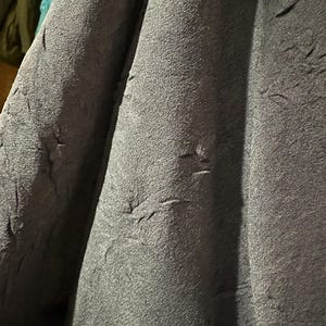 May include: Close-up of dark gray fabric with a textured, crinkled surface. The fabric appears to be draped, with folds and shadows adding depth. The material has a slightly rough, uneven texture.