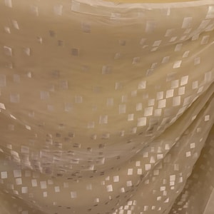 May include: Close-up of a flowing, sheer fabric in a pale yellow hue. The fabric is adorned with a pattern of small, square, reflective accents, creating a subtle, shimmering effect. The material drapes in soft folds, suggesting a light and airy texture.