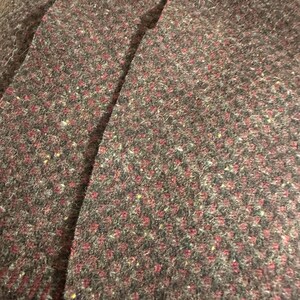May include: Close-up of a textured fabric with a speckled pattern. The fabric is primarily dark gray with small, evenly spaced red and yellow flecks. The material appears to be thick and possibly wool or a similar textile.