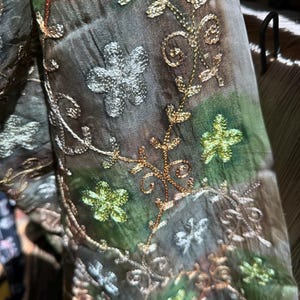 May include: A sheer fabric scarf featuring a patchwork of muted green and brown tones. The material is decorated with intricate embroidery in metallic gold, silver, and bright lime green threads, forming floral motifs and swirling vine patterns.