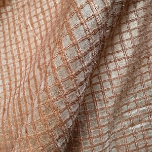 May include: Close-up of a textured fabric with a grid pattern. The fabric is a blend of peach and beige tones, with the grid lines in a darker peach hue. The fabric appears to be woven, with a slightly rough texture.