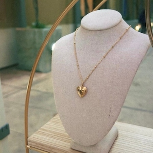 Puede incluir: Un collar de oro con un colgante en forma de corazón en un expositor de tela blanco. El collar tiene una cadena delicada y un dije de corazón detallado.