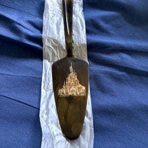 Puede incluir: Una espátula para pastel marrón oscuro con mango y una hoja ancha y plana. La hoja presenta una ilustración dorada y blanca de un castillo. La espátula descansa sobre papel blanco translúcido arrugado.
