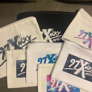 May include: A collection of zippered pouches in various sizes and colors. The pouches are white and black, with the text "WOXY FM 97.7" and "97X WOXY FM 97.7" printed on them in different colors and styles. The pouches are made of a canvas-like material.