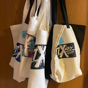 May include: Several white tote bags with black handles hang on a metal rack. The bags feature a colorful graphic design with the letters "TX" and the words "WXY FM 92.7". One bag has a blue sticky note with the number "#3".