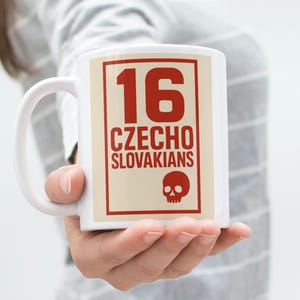 May include: White ceramic mug with a handle, featuring a red and beige graphic. The graphic includes the number "16", the words "CZECHO SLOVAKIANS", and a skull icon. The mug is held by a person.