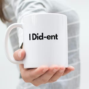 May include: White ceramic mug with the text "I Did-ent" in black. The mug has a curved handle and is held by a person. The background is a blurred, light gray and white.