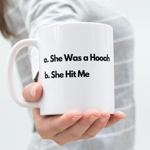May include: A white ceramic mug featuring black printed text that reads: a. She Was a Hooah, b. She Hit Me. The mug has a standard cylindrical shape with a curved handle on the left side, designed for holding hot or cold beverages in a simple style.