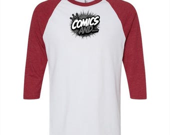 May include: A white baseball-style t-shirt with three-quarter length heather red sleeves. The center of the chest features a black and white graphic with a crown icon and the text "COMICS AND..." set against a circular, spattered ink background.
