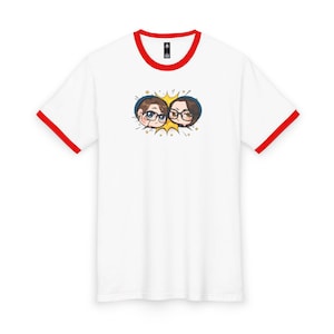May include: A white ringer t-shirt with red trim on the collar and sleeve cuffs. The center of the chest features a graphic illustration of two chibi-style characters with glasses, framed by a yellow starburst shape and small gold stars.
