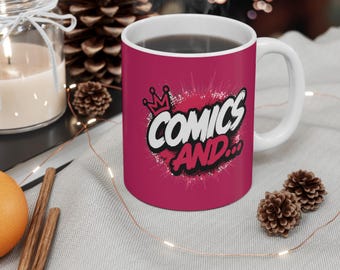 May include: A magenta ceramic mug with a white interior and handle, featuring a graphic design with a crown icon and the text "COMICS AND..." in bold white and black lettering, surrounded by a white splatter effect on the front surface.