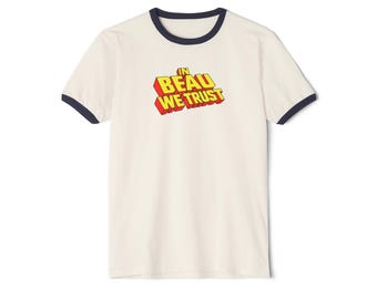 May include: A cream-colored ringer t-shirt with dark navy blue trim on the collar and sleeve cuffs. The center of the chest features the text IN BEAU WE TRUST in a bold, three-dimensional yellow font with a red outline and a drop shadow effect.