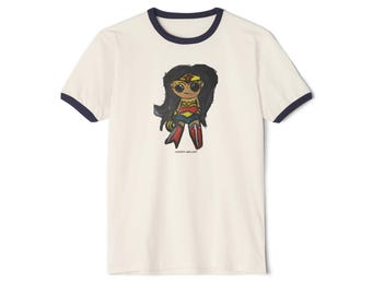 May include: A cream-colored ringer t-shirt with dark blue trim on the collar and sleeves. The center features a stylized, hand-drawn illustration of a character with long black hair, a gold tiara, and a red, blue, and gold outfit. Text reads COMIC-ART.COM.