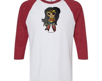 May include: A white baseball t-shirt with three-quarter length heather red sleeves. The front features a cartoon illustration of a character with long black hair, a gold tiara, a red and blue outfit, and red boots. The text COMICS-ART.COM is printed below.