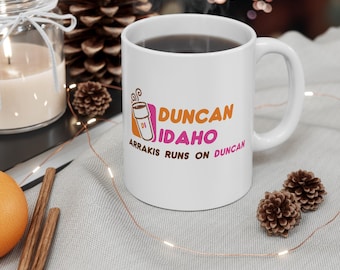 May include: A white ceramic mug featuring a graphic of a coffee cup and the text DUNCAN IDAHO with the slogan ARRAKIS RUNS ON DUNCAN. The design uses orange and pink lettering with a small coffee cup icon labeled DI on the left side.