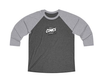 May include: A dark heather gray baseball shirt with light gray three-quarter length sleeves. The chest features a graphic with a small crown icon above the white text that reads COMICS AND... with a black shadow effect and a gray splatter background.