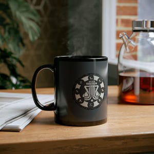 May include: A black ceramic mug with a white graphic design featuring a stylized emblem. The mug is on a wooden surface, with steam rising from it. A glass teapot with a dark liquid is in the background.