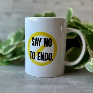 Puede incluir: Taza de cerámica blanca con un círculo amarillo y una línea diagonal que lo atraviesa. El texto "SAY NO TO ENDO." está impreso en negro dentro del círculo.