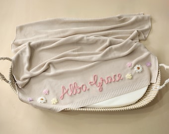 May include: A beige knitted baby blanket with the name "Alba Grace" embroidered in pink script. The blanket is decorated with small, colorful flower appliqués and is placed in a woven basket.