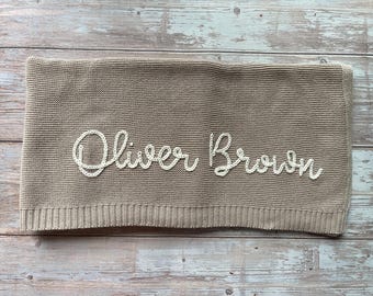 May include: A tan knitted blanket with the name "Oliver Brown" embroidered in white cursive script. The blanket has a ribbed border and is folded neatly. The background is a wooden surface.