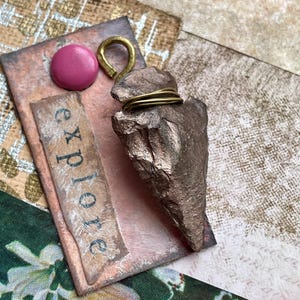 May include: A handmade pendant featuring a bronze-colored arrowhead wrapped with gold wire. The pendant is attached to a pink and copper-colored rectangular base with the word "explore" printed on it. A pink button and gold hook are also attached.