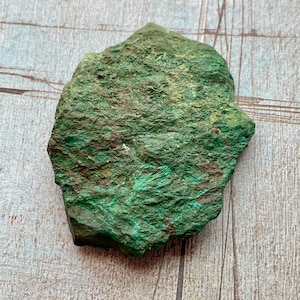 May include: A rough-hewn, irregularly shaped, green mineral specimen. The surface has a textured appearance with variations in color, ranging from deep emerald to lighter shades of green. Small patches of brown are visible.