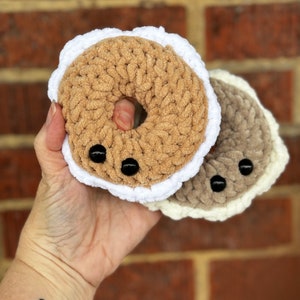Puede incluir: Dos juguetes de peluche con forma de donut hechos a crochet. Uno es marrón con ribete blanco y el otro es gris con ribete blanco. Ambos tienen ojos de botón negros.