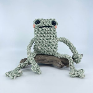 May include: A green crocheted frog toy sitting on a piece of driftwood. The frog has black eyes and pink cheeks.