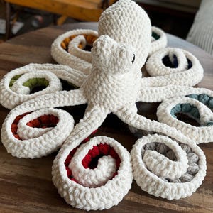 May include: A handmade, plush octopus toy in cream-colored yarn. The octopus has eight curled tentacles with colorful interiors in red, green, blue, gray, and orange. The toy is sitting on a wooden surface.