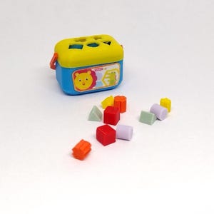 May include: A blue and yellow plastic toy box with a lion face on the front. The box is open and there are various colored plastic shapes inside, including squares, triangles, and cylinders.