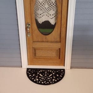 May include: A black metal half-round doormat with a decorative, intricate design. The doormat is placed in front of a wooden door with a glass oval window.