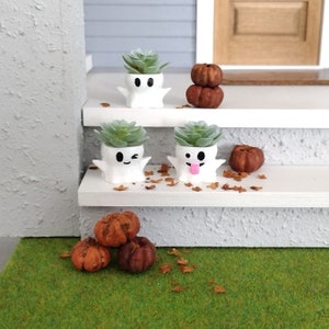 May include: Three white ghost-shaped planters with cute faces, each holding a small green succulent plant. The planters are arranged on a white shelf with two small orange pumpkins on the bottom shelf.
