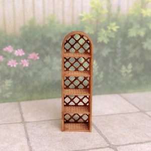 May include: A miniature wooden shelf unit with an arched top and open lattice-style sides. The shelves are rectangular and the unit is a light brown color. The background is a blurred outdoor scene with greenery and a fence.