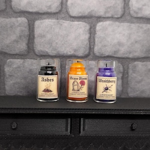 May include: Three small glass jar candles with labels reading "Ashes", "Grave Bloom", and "Wraithberry". The candles are black, orange, and purple. The labels have illustrations and text. The candles are on a black table.