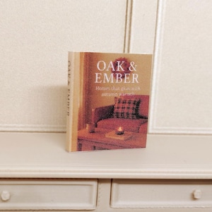 May include: A hardcover book titled "OAK & EMBER" with a cover image of a cozy living room scene. The book is standing upright on a white surface, with the title and author's name clearly visible. The book's spine also displays the title.