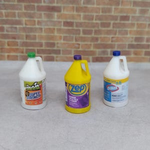 May include: Three miniature cleaning product bottles. One is white with a green cap and orange label, another is yellow with a purple label, and the third is white with a blue cap and label. The bottles have handles.