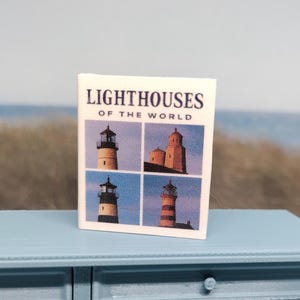 May include: A miniature book titled "Lighthouses of the World" featuring four lighthouse images. The book is on a light blue table with a scalloped edge, set against a blurred background of a beach and ocean.
